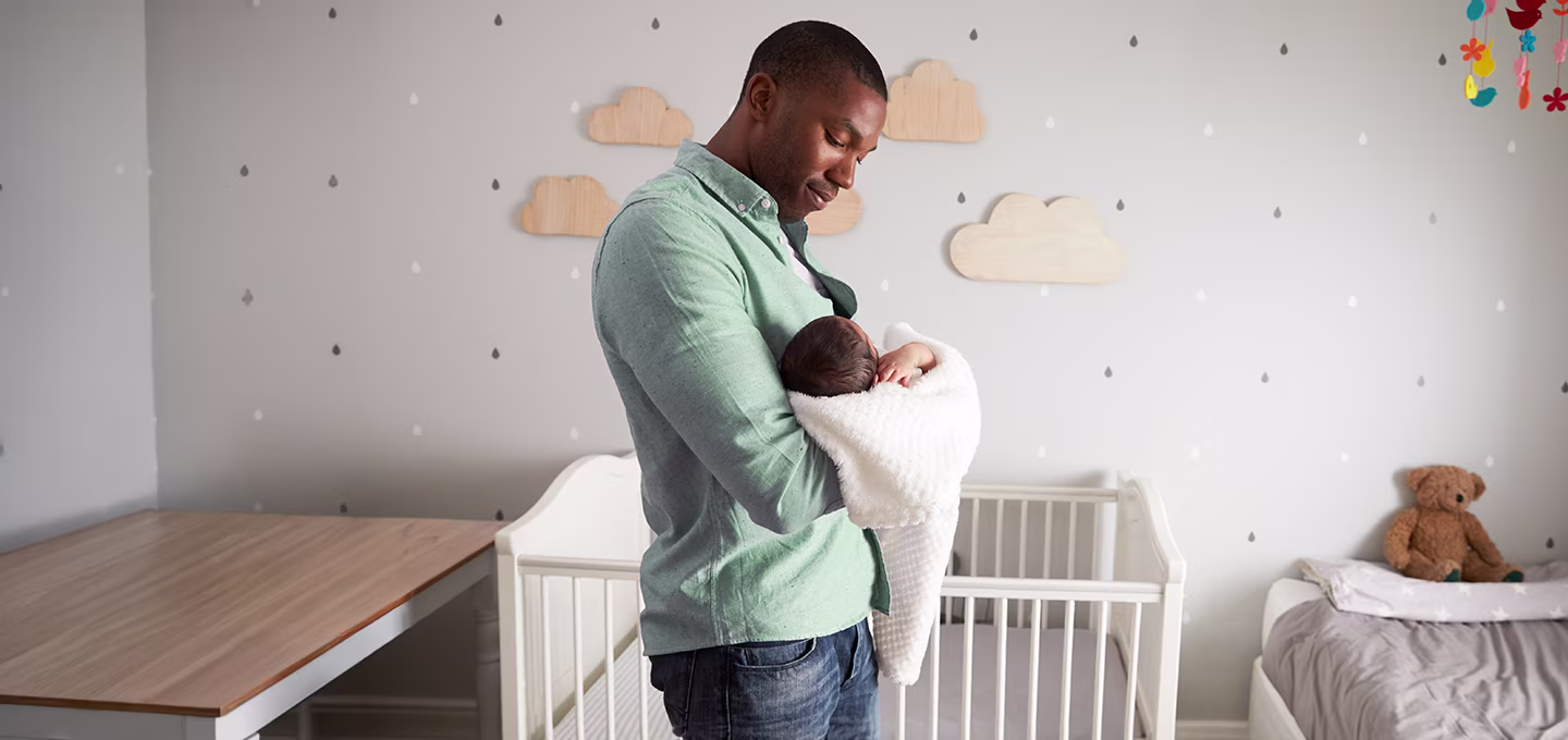 Father lovingly holding newborn in a cozy nursery with cloud decor and crib.