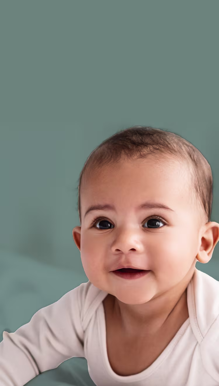 Close up of baby laying on stomach smiling and wearing a cream onesie with sage background