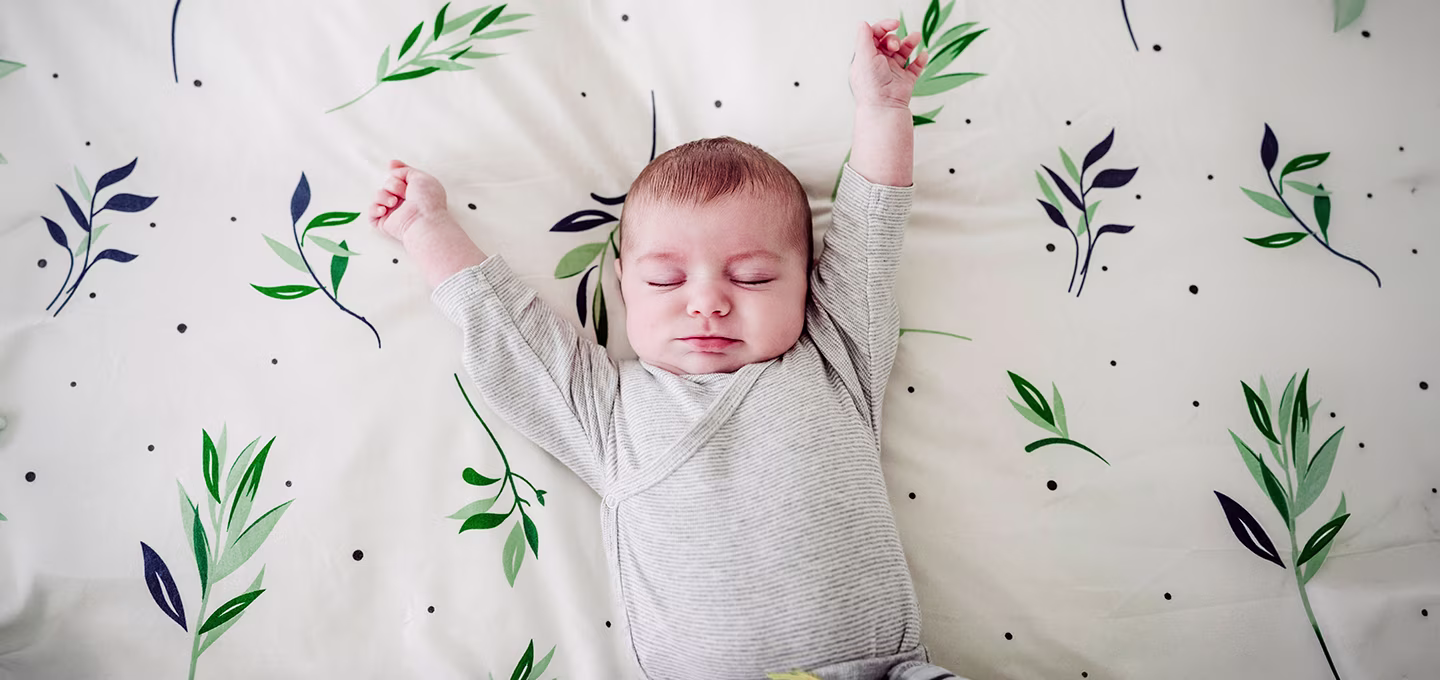 Baby sleeping peacefully on a patterned sheet, wearing a grey onesie.