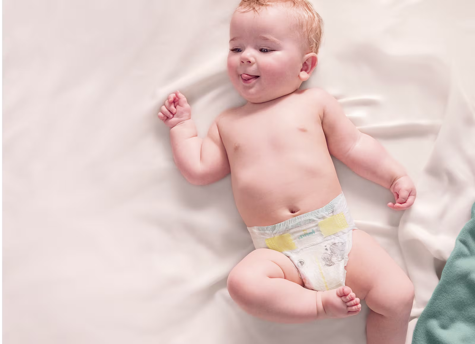 Ein Baby in einer Windel liegt auf einer weichen, weißen Decke, lächelt und wirkt verspielt, während es die Zunge herausstreckt, und vermittelt so ein Gefühl von Freude und Unschuld.