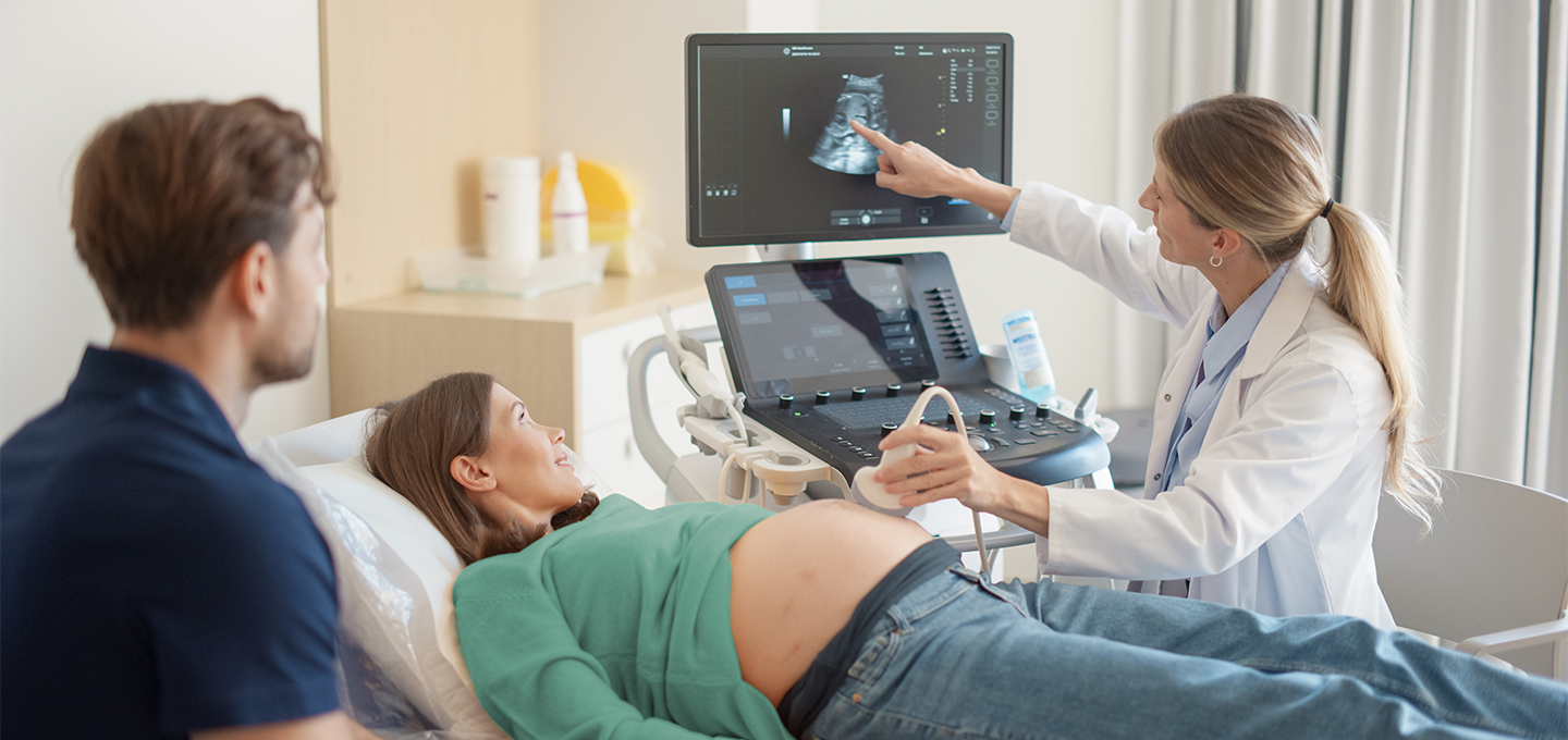 A pregnant woman lies on an exam table while a female ultrasound technician points to a monitor showing a sonogram of the fetus.