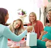 Women at a baby shower, exchanging gifts and smiling with balloons in the background.
