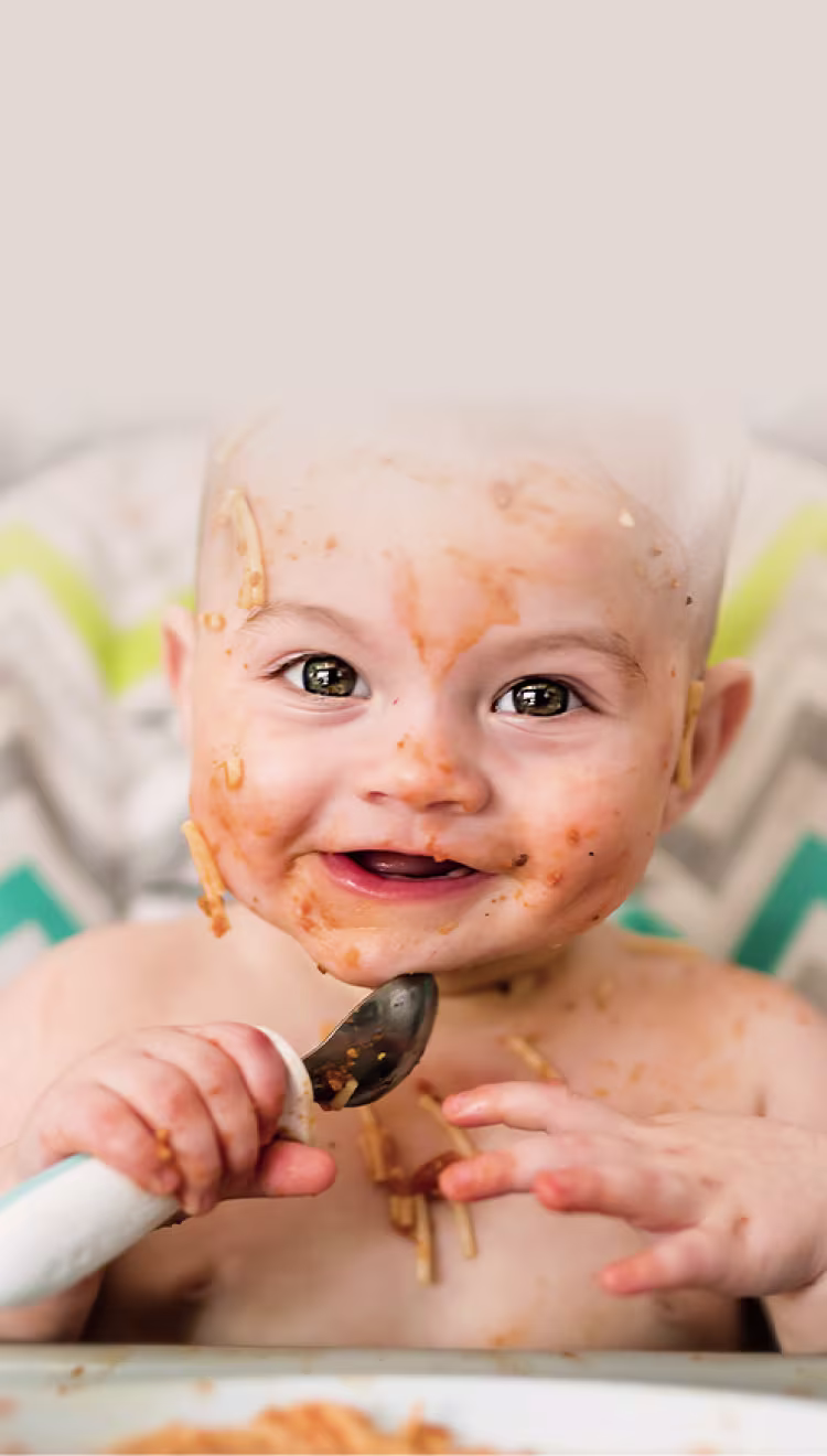 Bebé sonriente en una trona, con la cara cubierta de salsa de espagueti, sosteniendo una cuchara