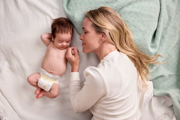 Uma mãe deitada numa cama segura ternamente a mão do seu recém-nascido de fralda, ambos tranquilos e próximos um do outro.