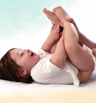 Bebelușul stă întins pe spate pe o suprafață albă, ținând picioarele ridicate, în timp ce părintele folosește un șervețel pentru a curăța fundul copilului.