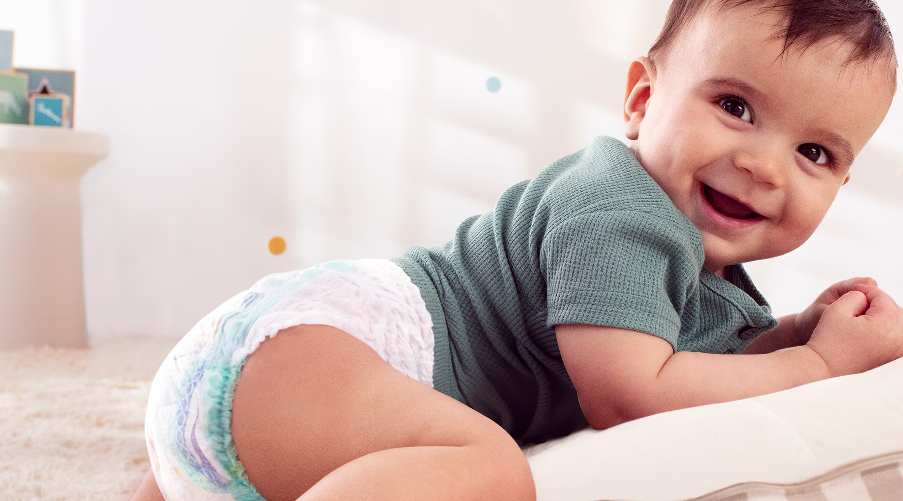 Een baby met een luier lacht naar de camera terwijl hij op zijn buik op een gestreept kussen ligt.