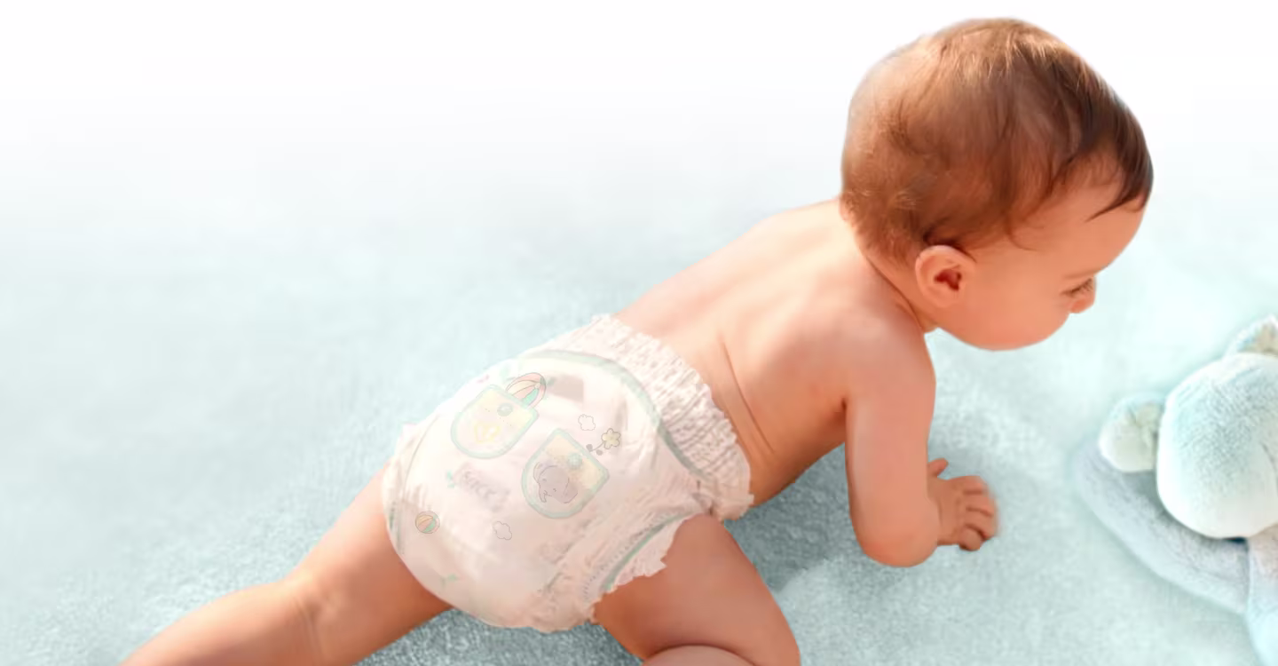 Een baby in luiers kruipt over een zachte ondergrond, naast hem ligt een knuffel, en hij toont zich speels en nieuwsgierig.