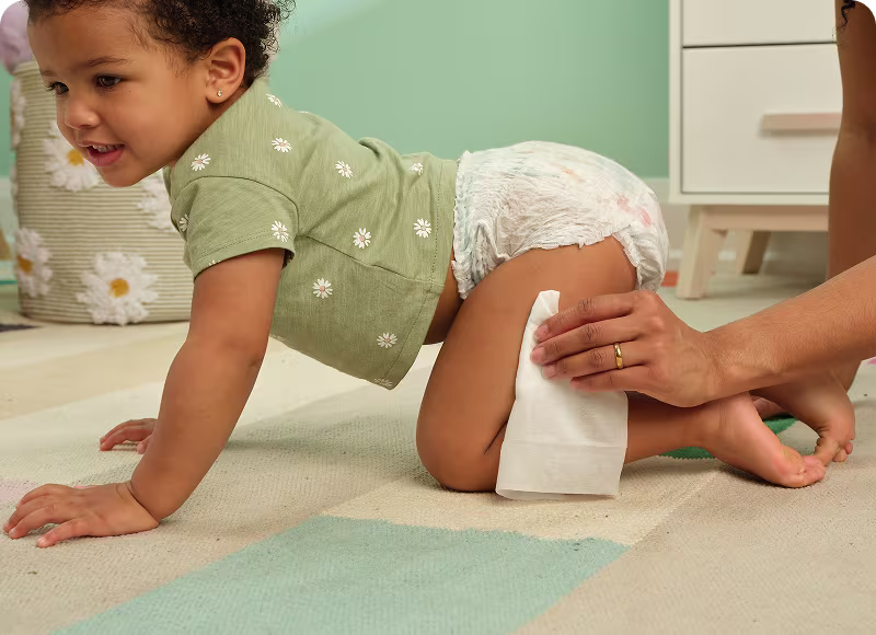 Mamá limpia la pierna de su bebé que gatea mientras exploran el cuarto, el bebé lleva un pañal tipo pull-up de Pampers y una camiseta verde con margaritas blancas.