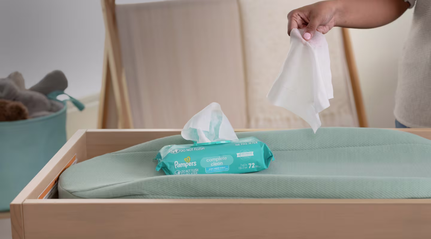 Una madre sacando una sola toallita Complete Clean del paquete que descansa sobre el colchón del cambiador, mostrando cómo está diseñado para funcionar la tapa superior de un solo pop de toallita.
