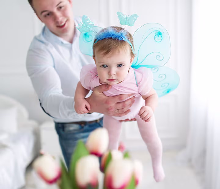 Baby in butterfly costume held by adult in a bright room with flowers in the foreground.