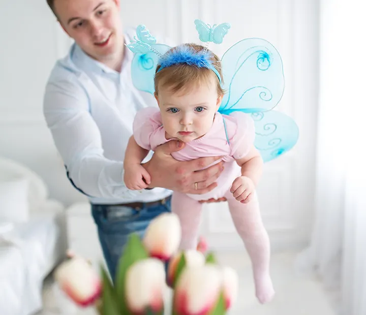 Baby in butterfly costume held by adult in a bright room with flowers in the foreground.