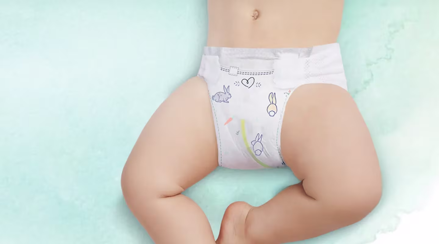 Fotografia arată partea inferioară a trunchiului și picioarele unui bebeluș întins pe o suprafață de culoare verde deschis, purtând un scutec Pampers cu indicatorul de umiditate vizibil.