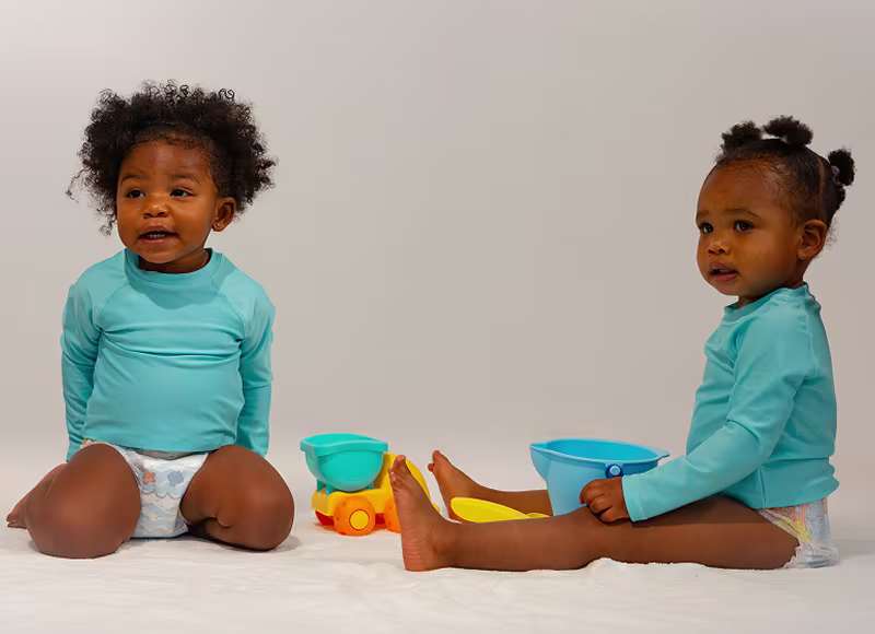 Dos niños pequeños, vestidos con bañadores Dodot Splashers y trajes de baño turquesa, están sentados jugando con una pala y un cubo.