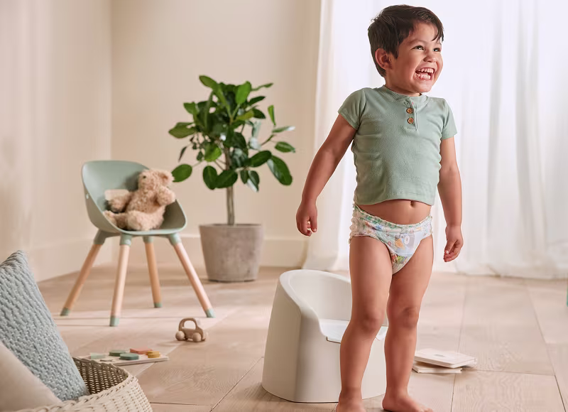 A smiling toddler wearing a Pampers Easy Ups training pant and sage green shirt stands in a bright living room with a plant, 