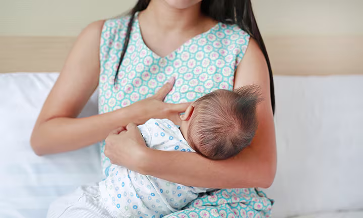 Foto de una madre amamantando a su bebé mientras está sentada en la cama, ilustrando consejos para la lactancia materna y una posición adecuada al dar el pecho.