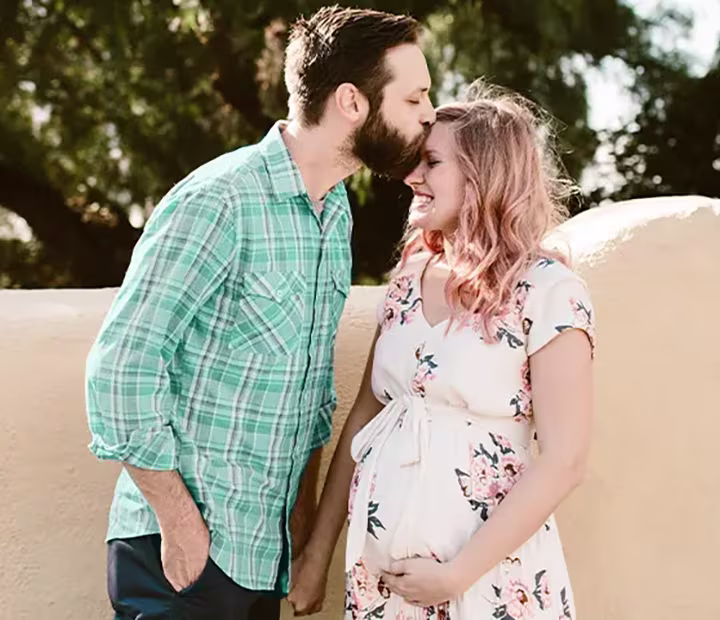 Expectant couple sharing a tender moment outdoors, with man kissing woman's forehead.