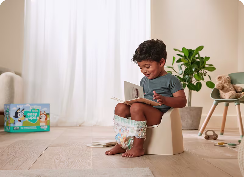 A toddler sitting on a potty reading a book, wearing Easy Ups