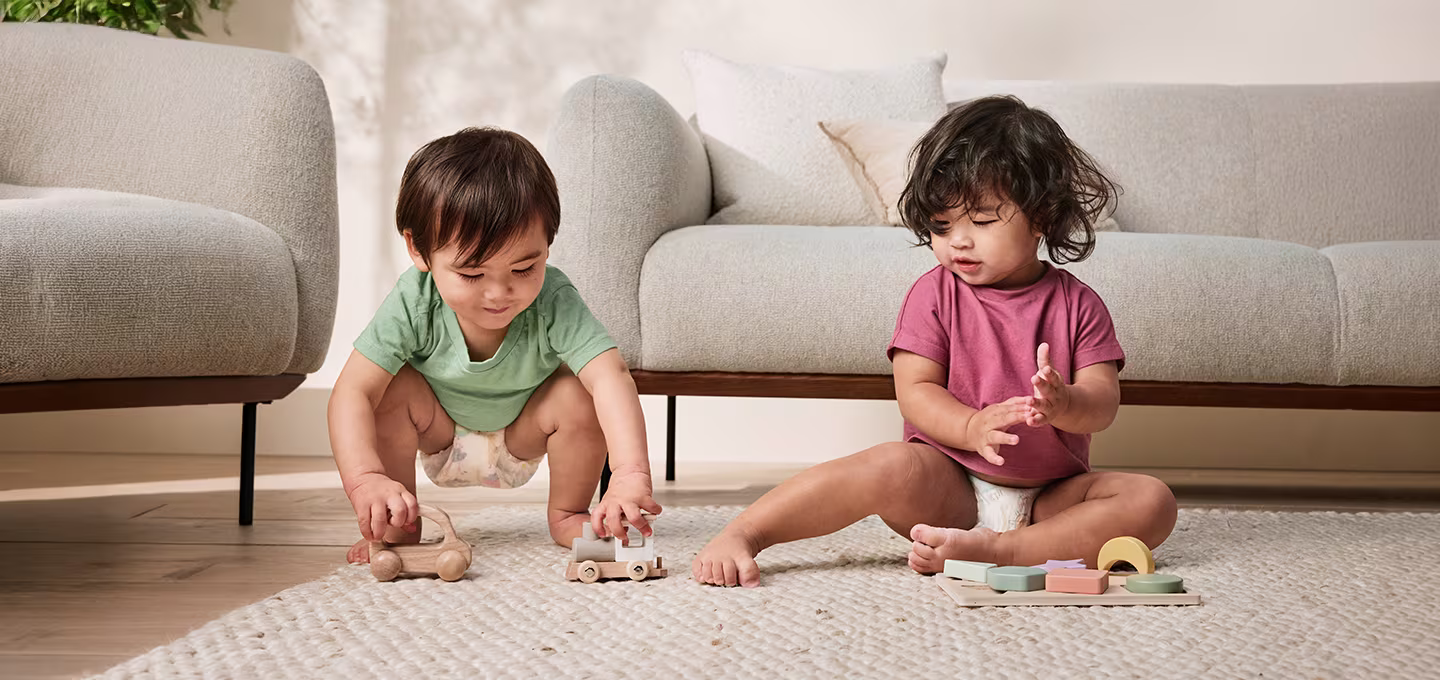 Zwei Kleinkinder in Windeln spielen mit Holzspielzeug auf einem Teppich in einem hellen Wohnzimmer.