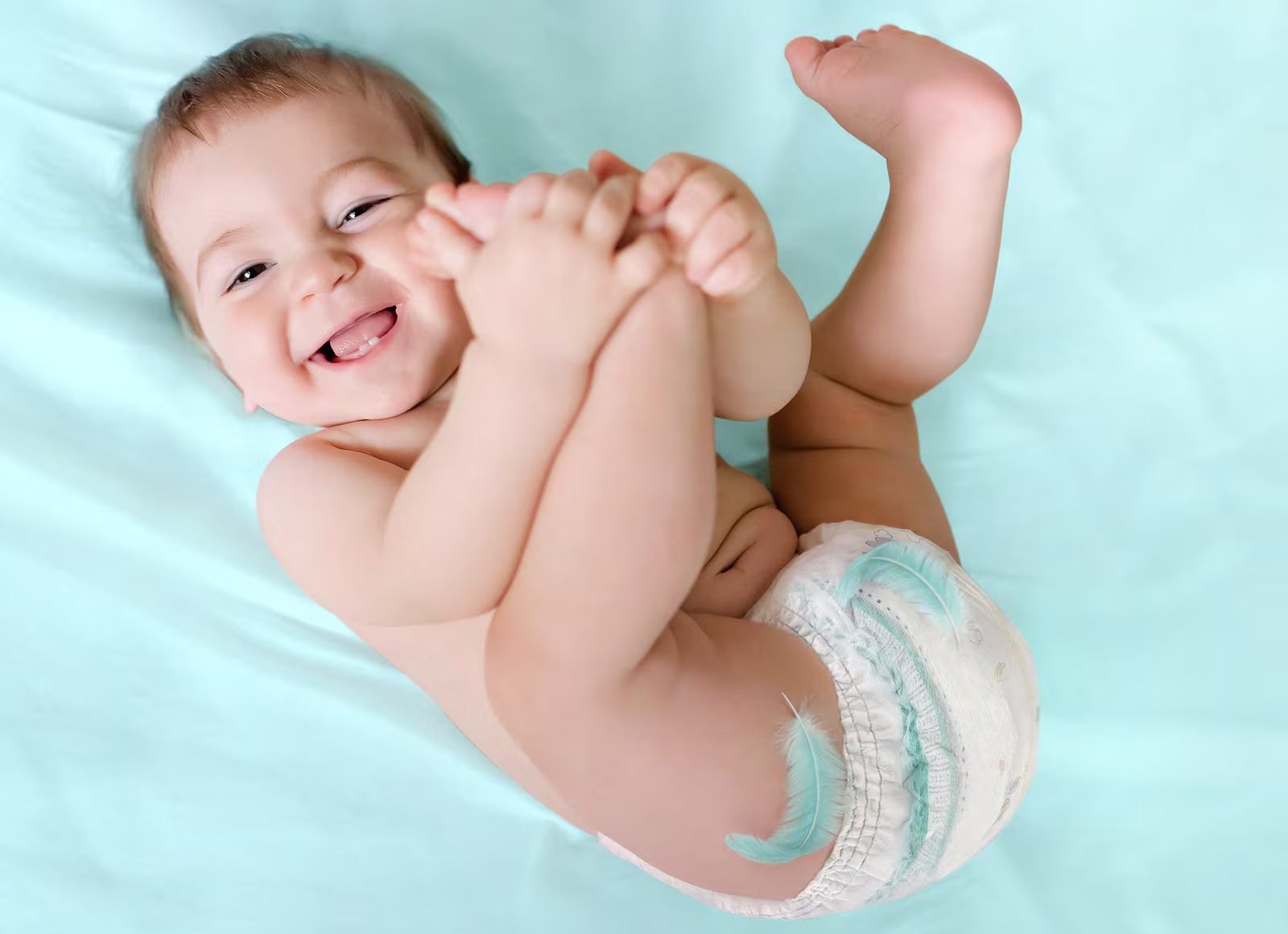 Ein glückliches Baby liegt auf dem Rücken auf einem hellblauen Laken, hält seine Füße und trägt eine Pampers® Premium Protection™ Pants