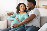 Expecting couple smiling, sitting together on a couch, with dad's hand on mom's baby bump.