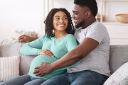 Expecting couple smiling, sitting together on a couch, with dad's hand on mom's baby bump.
