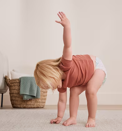 A playful toddler in a red shirt doing a triangle yoga pose in her room, bending down with one hand on the ground and other up towards the sky, showing Swaddlers 360 can move with your baby