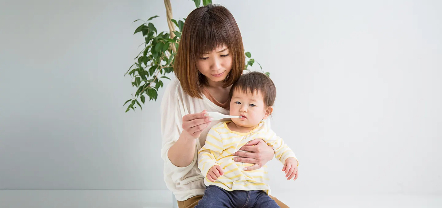 ES Baby Diarrhea - Hero image - Dec 2025 Madre sentada sostiene a su bebé mientras le ofrece una cucharada de líquido, ilustrando cuidado y alimentación durante un episodio de diarrea infantil.