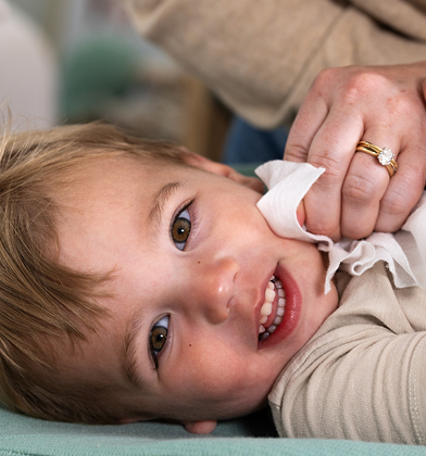 A toddler with brown hair and light brown eyes looks up from a green changing pad, smiling while a woman's hands gently wipe their face with a white wipe.