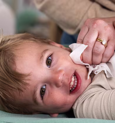A toddler with brown hair and light brown eyes looks up from a green changing pad, smiling while a woman's hands gently wipe their face with a white wipe. A toddler with brown hair and light brown eyes looks up from a green changing pad, smiling while a woman's hands gently wipe their face with a white wipe.