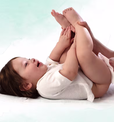 Niemowlę leży na plecach na białej powierzchni, trzymając stopy w górze, podczas gdy rodzic używa chusteczki do wyczyszczenia pupy dziecka.