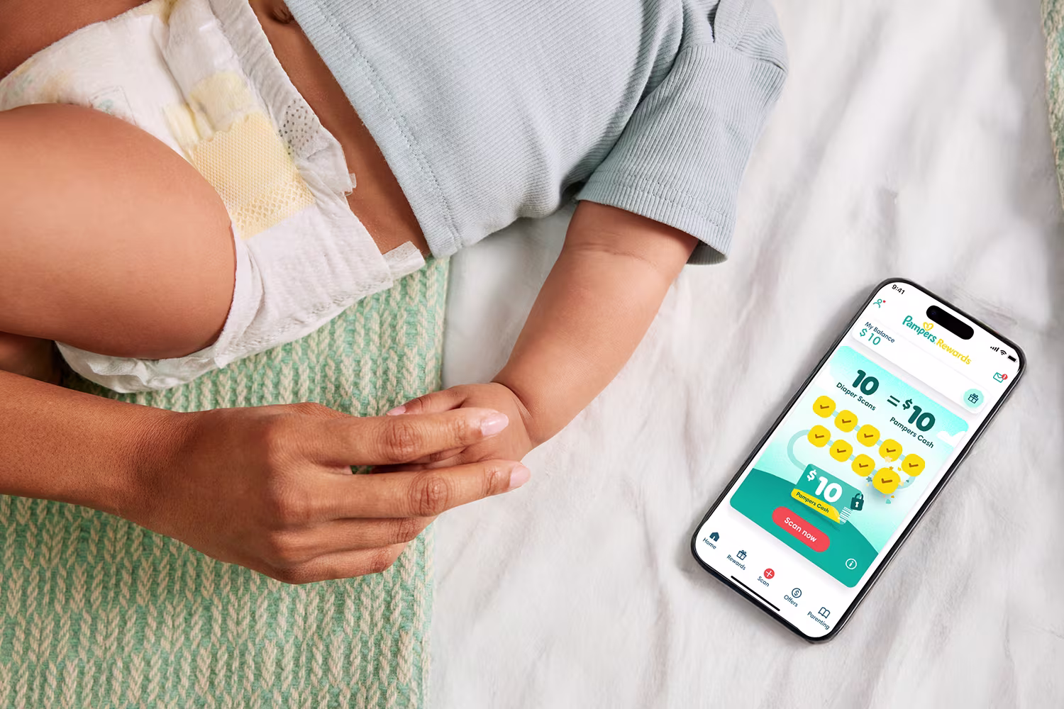 A close-up shows an adult’s hand gently holding a baby’s forearm while the baby lies on a bed wearing a diaper and a light-colored top. Next to them on the bed is a smartphone displaying the Pampers reward app, and showcasing that 10 scans on the app equals 10 dollars rewards. 