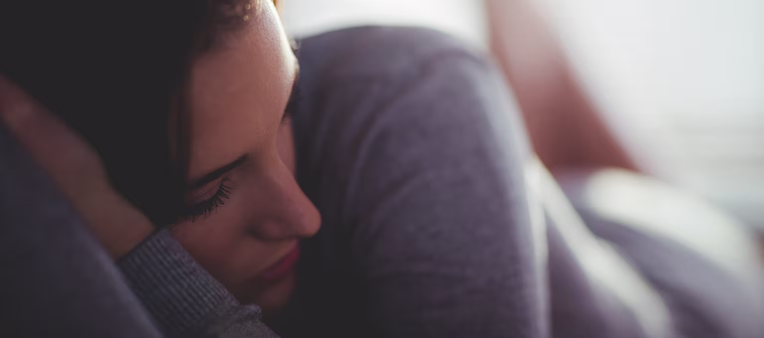 Close-up of a woman lying on her side with her head resting on her arm, looking down with a withdrawn expression, representing postpartum depression.