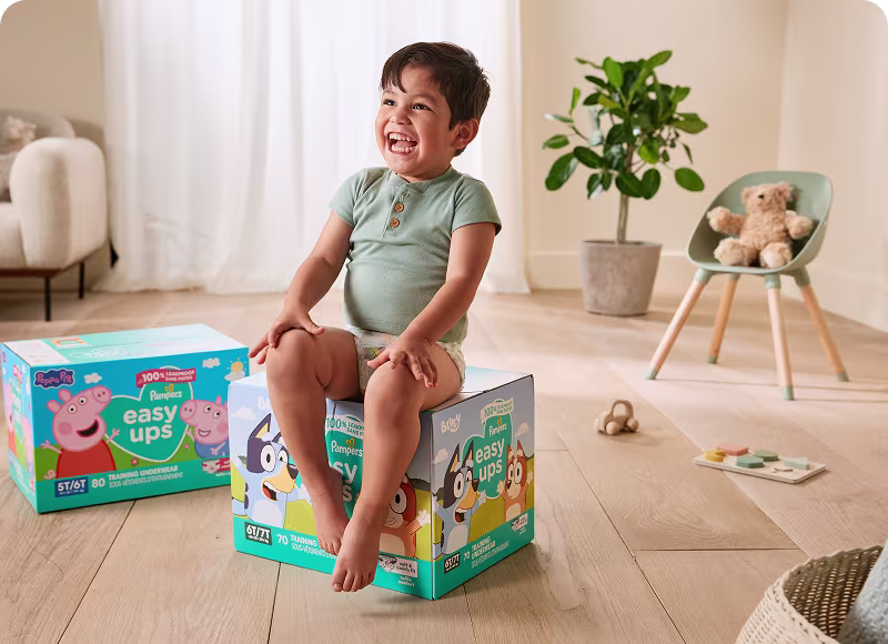 A laughing toddler sitting on a box of Easy Ups in the kiving room with another box, chair and plant in the background