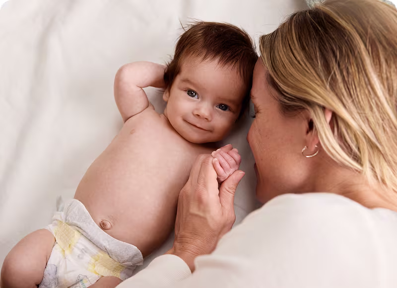 Imagen que muestra a un bebé sonriendo boca arriba con Pampers Swaddlers y su mamá a la derecha sosteniendo su mano Imagen que muestra a un bebé sonriendo boca arriba con Pampers Swaddlers y su mamá a la derecha sosteniendo su mano