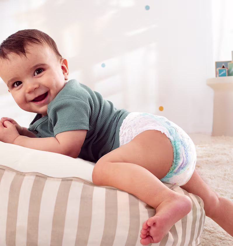 Een baby met een pants-luier lacht breed naar de camera terwijl hij op zijn buik op een gestreept kussen ligt. Een baby met een pants-luier lacht breed naar de camera terwijl hij op zijn buik op een gestreept kussen ligt.