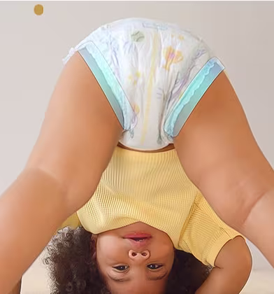 Małe dziecko w pieluszce jest pokazane do góry nogami, gdy patrzy na kamerę między swoimi nogami, wykonując odwróconą pozę na jasnym dywanie.