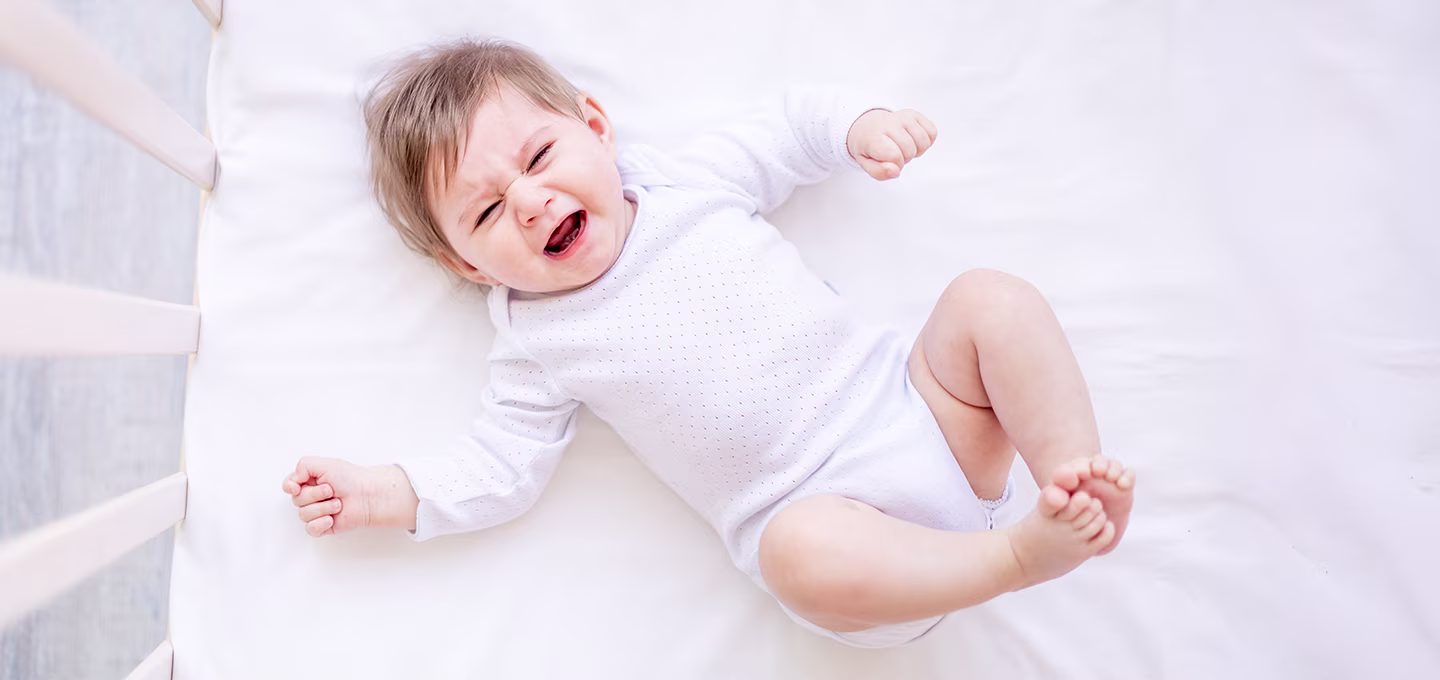 Bebé acostado en cuna llorando con expresión de malestar, piernas y brazos tensos, ilustrando síntomas típicos de cólico infantil.