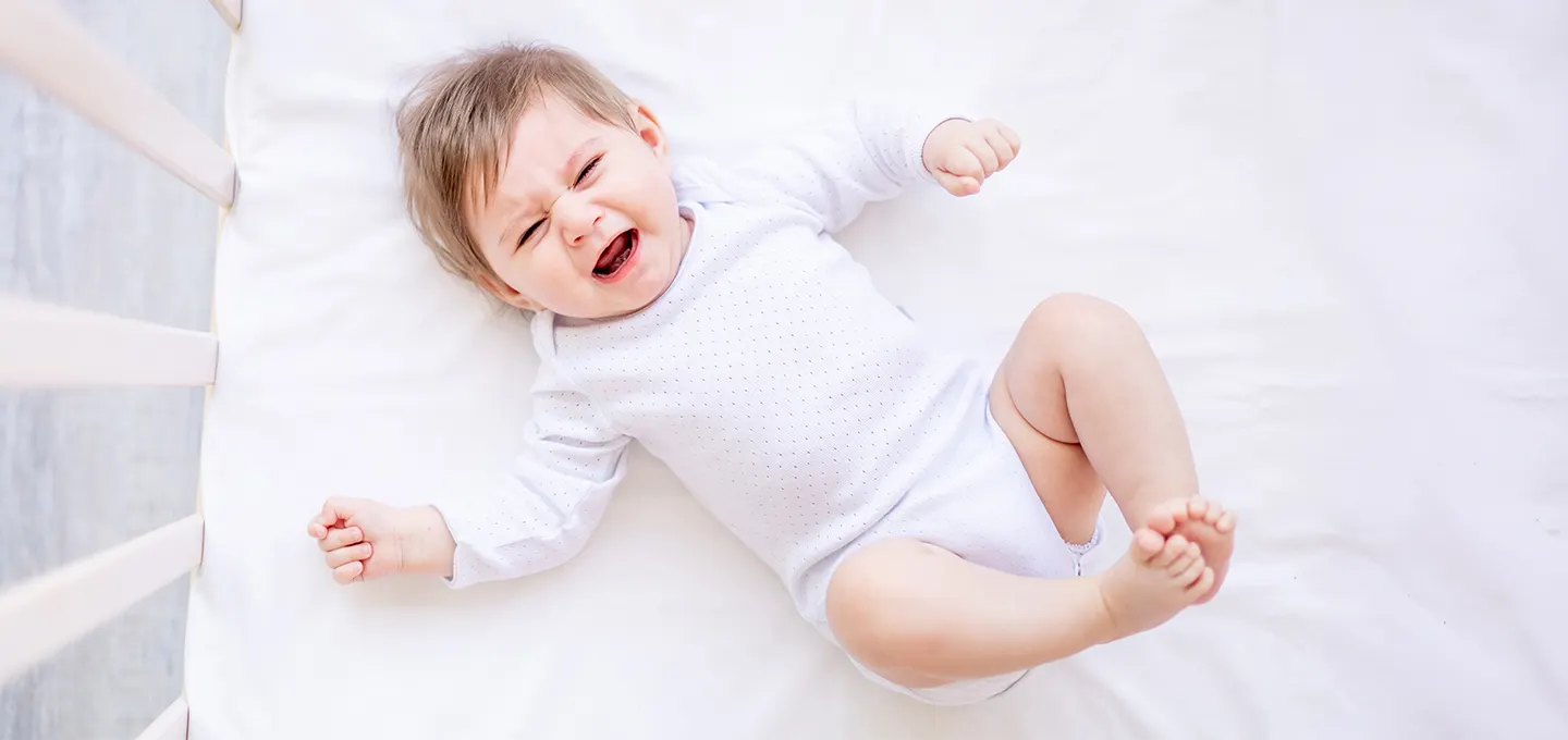 Bebé acostado en cuna llorando con expresión de malestar, piernas y brazos tensos, ilustrando síntomas típicos de cólico infantil.