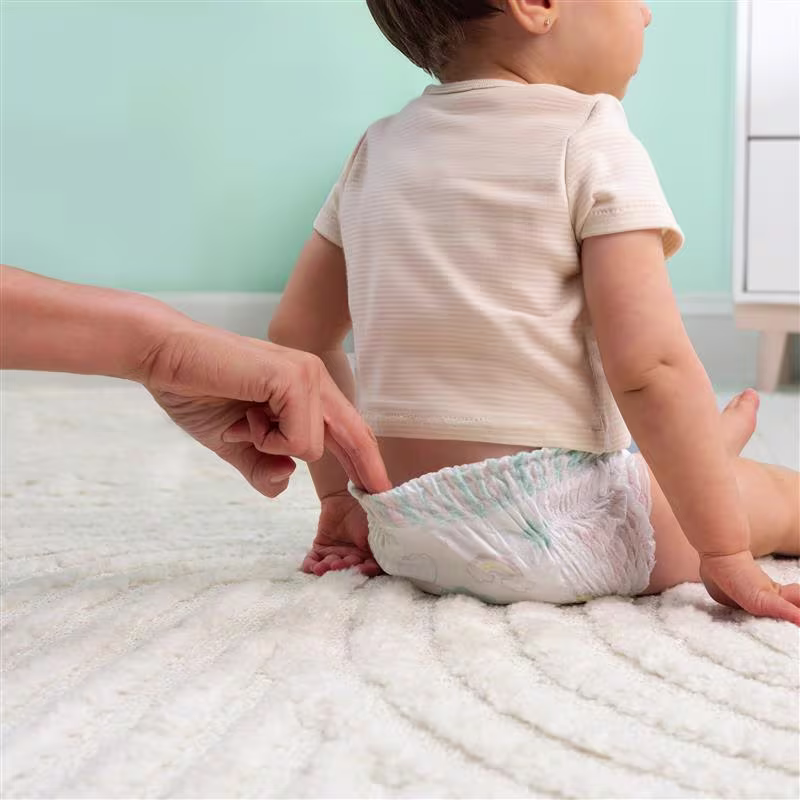 Un padre jalando suavemente la parte trasera de un pañal Swaddlers 360 pull on mientras el bebé está sentado en su cuarto de bebé. Un padre jalando suavemente la parte trasera de un pañal Swaddlers 360 pull on mientras el bebé está sentado en su cuarto de bebé.