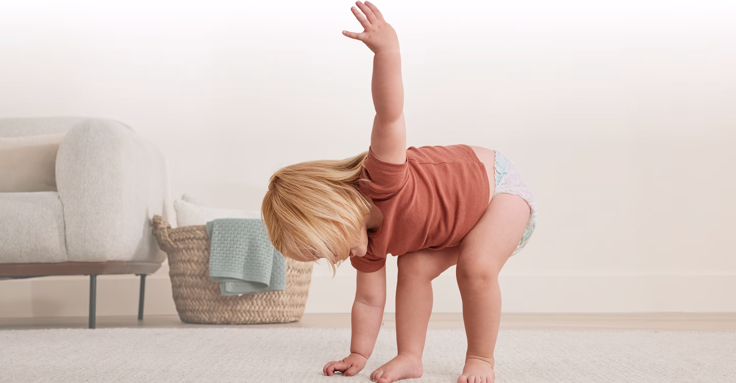 Małe dziecko chodzi po jasnym dywanie obok szarej sofy.