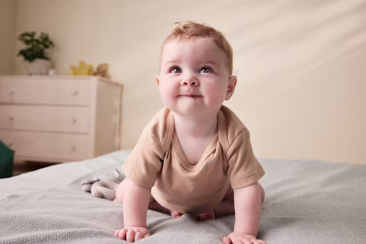 Um bebé em body bege, de gatinhas sobre uma cama, olha para cima com uma expressão curiosa e alegre.