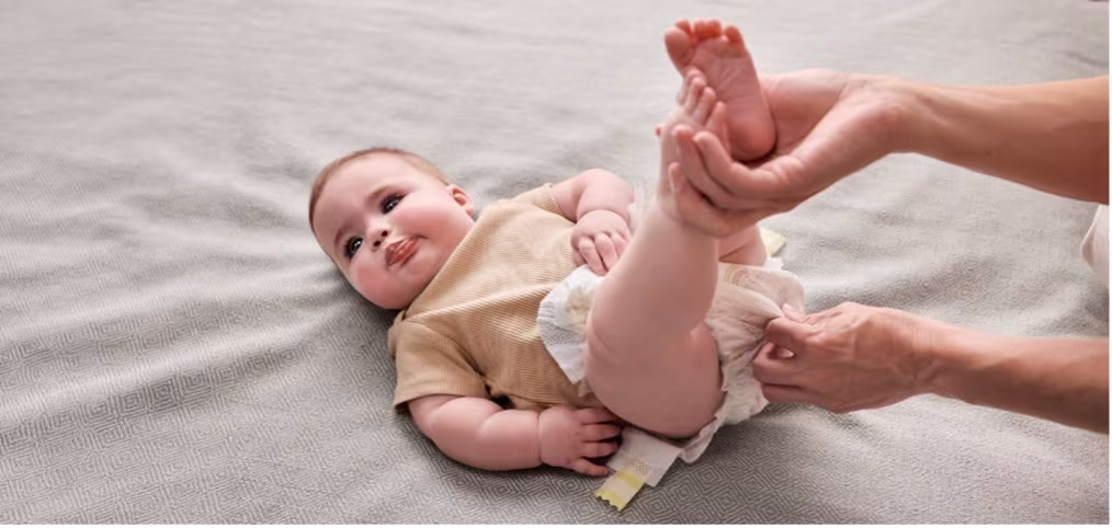 Un adulte change la couche d'un bébé tout en lui tenant délicatement les jambes, sur une surface moelleuse.