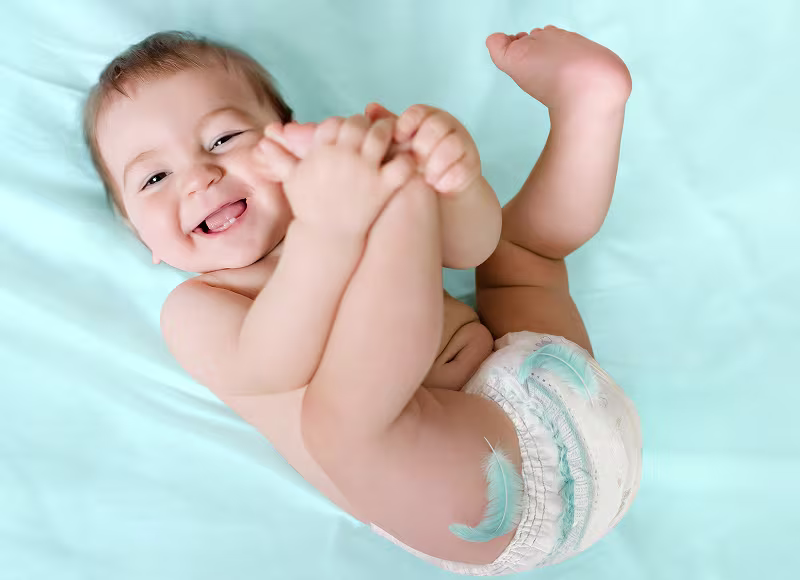 Szczęśliwe dziecko w pieluszce Pampers, leżące na jasnoniebieskim prześcieradle i trzymające swoje stopy.