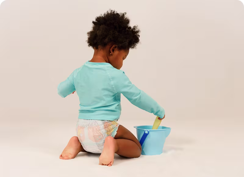 Un niño pequeño con Pampers Splashers y una camiseta de baño color teal se arrodilla mientras juega con una pala y un balde.