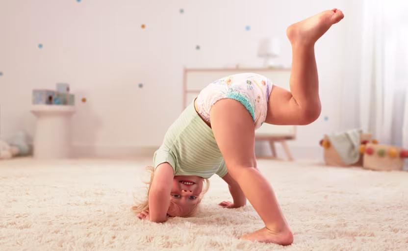 Ein glücklicher Kleinkind in Pampers Baby-Dry Pants macht einen Handstand auf einem flauschigen Teppich und lächelt in die Kamera.