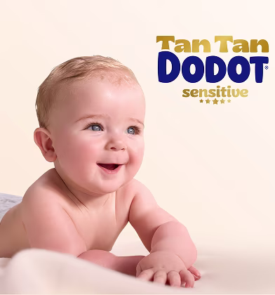 Um bebé feliz com olhos azuis e cabelo claro sorri enquanto está deitado de barriga para baixo, usando uma fralda Dodot, olhando para a esquerda.