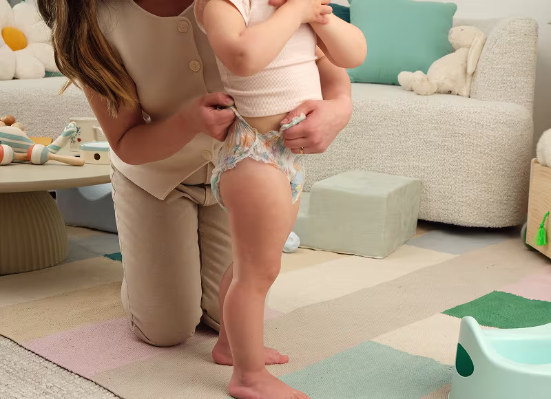 Close up of a toddler wearing Easy Ups, standing in a living room with mom kneeling beside her, carefully tearing the sides of her Easy Ups training pant. A couch, coffee table and potty in the background 