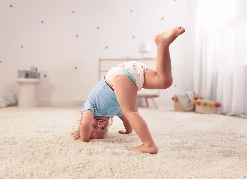 Un copil fericit în scutec Pampers Active Baby se apleacă și se sprijină pe mâini pe un covor pufos, zâmbind către cameră. Un copil fericit în scutec Pampers Active Baby se apleacă și se sprijină pe mâini pe un covor pufos, zâmbind către cameră.