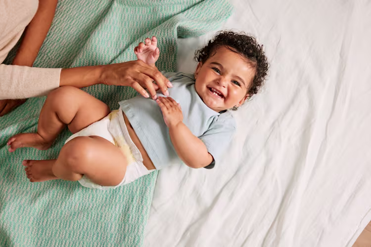 Um bebé sorridente deitado numa manta, a brincar enquanto um dos pais lhe segura suavemente na mão.
