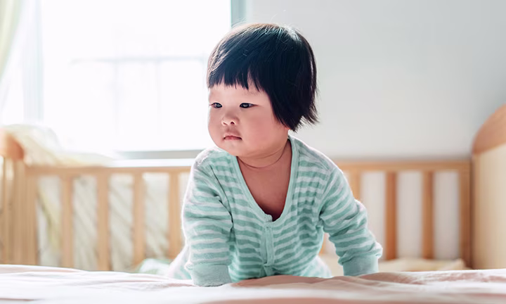 Baby in striped onesie crawling on a bed in a bright room.